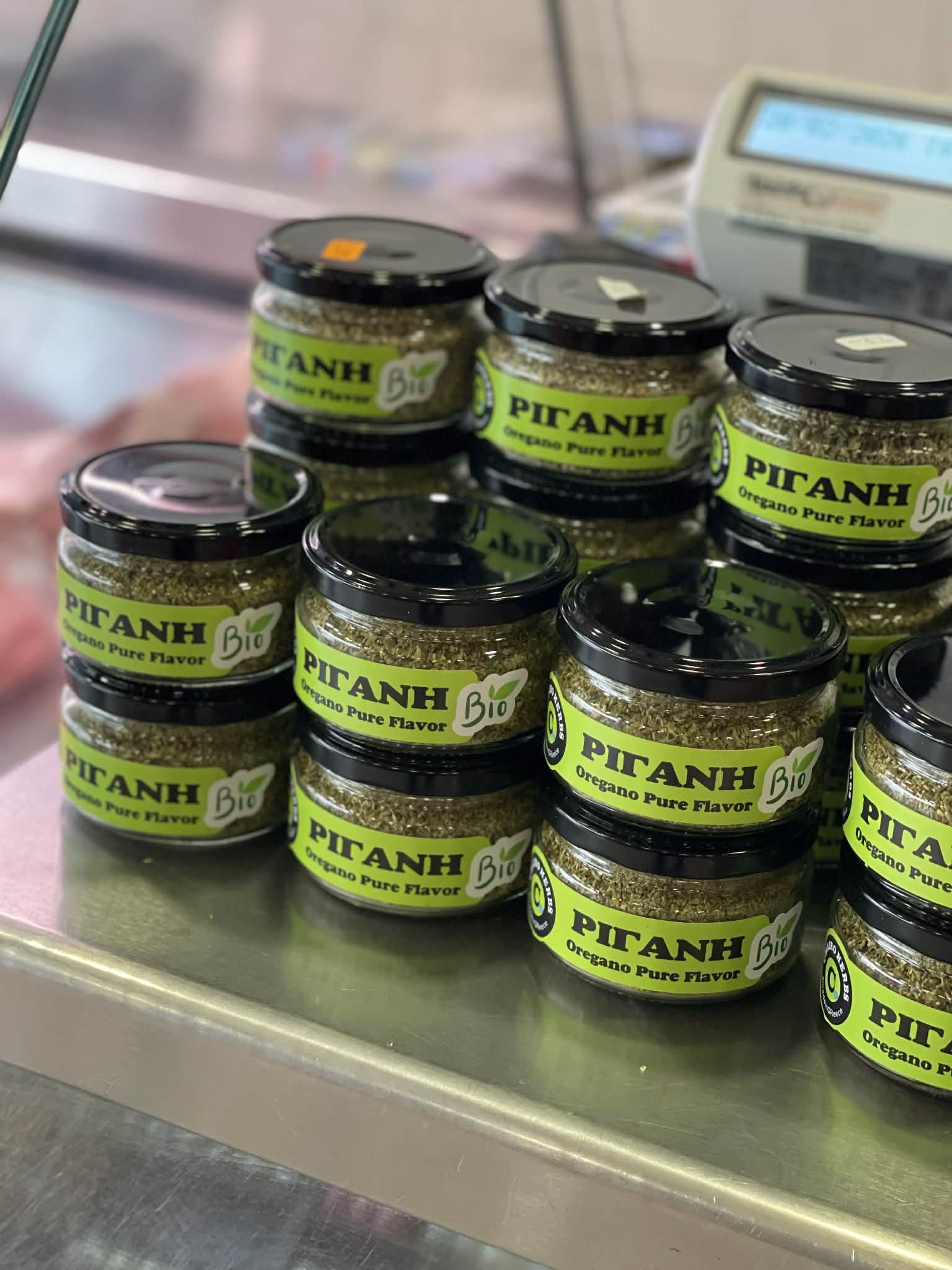Stacks of small glass jars containing dried oregano on a stainless steel shop counter.