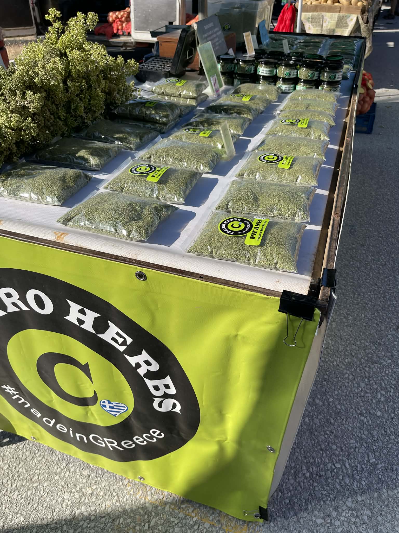 Packaged dried herbs and jars displayed on a market stall with a green Greek banner.